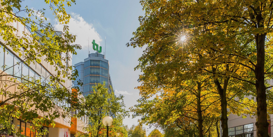 Rechts und links Bäume in herbstlichen rot-brauen Farben und ein TU Dortmund Gebäude mit Logo in der Mitte.