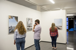 Drei Personen schauen sich Fotografien an, die an einer weißen Wand hängen.