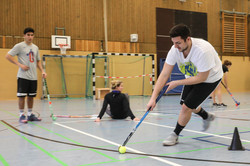 Zwei Studenten spielen Hockey, eine Studentin sitzt auf dem Boden der Turnhalle