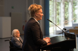 Profil ab der Hüfte von Wissenschaftsministerin Ina Brandes, eine Frau mit blonder Hochsteckfrisur und schwarzem Blazer. Sie steht an einem Rednerpult und spricht. Im Hintergrund sitzt Horst Selbiger, ein älterer Mann mit grauem Zopf und Brille, und hört zu.