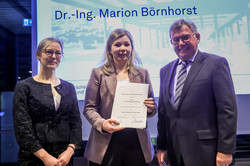 Gruppenfoto: Drei Personen stehen vor einer Leinwand, eine davon hält eine Urkunde.