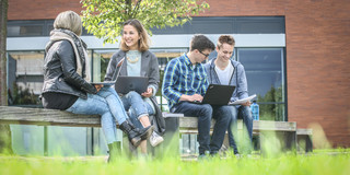 Studierende auf dem Campus Auf einer Bank vor einem Uni-Gebäude sitzen vier Studierende und unterhalten sich