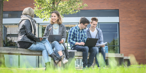 Auf einer Bank vor einem Uni-Gebäude sitzen vier Studierende und unterhalten sich