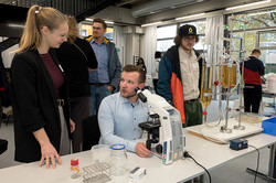 Das Bild zeigt Personen vor einer Tischtafel, auf der verschiedene Modelle stehen. Eine Person sitzt vor einem Mikroskop.