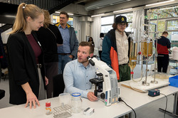 The picture shows people in front of a table with various models on it. One person is sitting in front of a microscope.