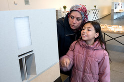 The picture shows a child in a pink jacket with a woman leaning down towards him. Both are looking at a white picture of a building. In the background is a table with a wooden model of the city.