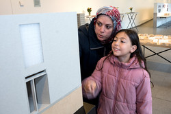 Das Bild zeigt ein Kind in einer rosafarbenen Jacke, zu dessen sich eine Frau runterbeugt. Beide schauen sich ein weißes Gebäudebild an. Im Hintergrund steht ein Tisch mit einem Stadtmodell aus Holz.