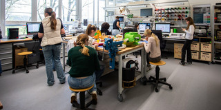 Foto: Mehrere Personen arbeiten gemeinsam in einer Werkstatt mit 3D-Druckern, Computern und verschiedenen Materialien.