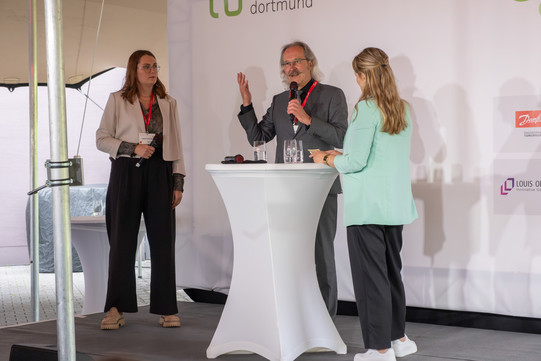 Begrüßung Fakultätsmitglieder Drei Personen stehen auf einer Bühne rund um einen Stehtisch.