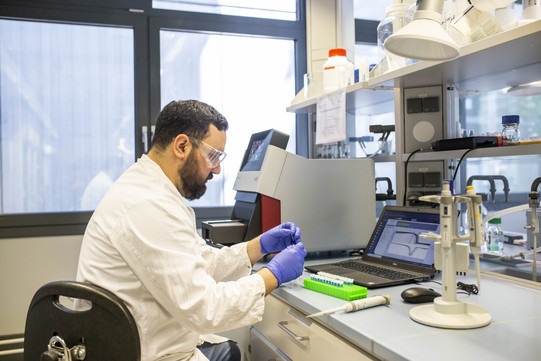 The photo shows Mr. Nicolás Gajardo-Parra sitting in the laboratory with protective clothing. In front of him is, among other things, an opened laptop.