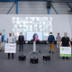 Gruppenbild mit mehreren Personen, im Hintergrund eine Leinwand mit Teilnehmern von Videochats