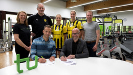 Zwei Männer sitzen an einem Tisch, auf dem ein TU-Logo steht und schreiben auf ein Stück Papier. Hinter ihnen stehen zwei Frauen mit einem Trikot von Borussia Dortmund sowie zwei Männer und eine weitere Frau. Im Hintergrund sind Fitnessgeräte zu erkennen. 