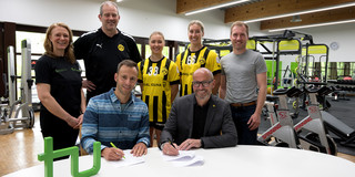 Zwei Männer sitzen an einem Tisch, auf dem ein TU-Logo steht und schreiben auf ein Stück Papier. Hinter ihnen stehen zwei Frauen mit einem Trikot von Borussia Dortmund sowie zwei Männer und eine weitere Frau. Im Hintergrund sind Fitnessgeräte zu erkennen. 