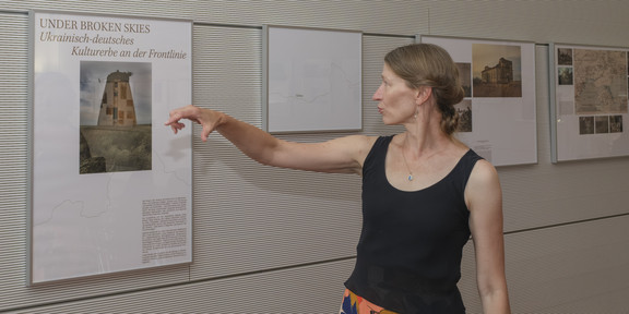 Foto: Eine Person zeigt auf ein Ausstellungsplakat an einer Wand mit Fotografien.