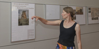 Foto: Eine Person zeigt auf ein Ausstellungsplakat an einer Wand mit Fotografien.