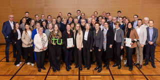 Group photo of the scholarship holders and donors in front of a wooden wall. In the front row, a young woman holds a green TU logo in her hands, two other women hold certificates.