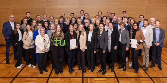 Gruppenfoto der Stipendiat*innen und Stifter*innen vor einer Holzwand. In der ersten Reihe hält eine junge Frau ein grünes TU-Logo in den Händen, zwei andere Frauen halten Urkunden.