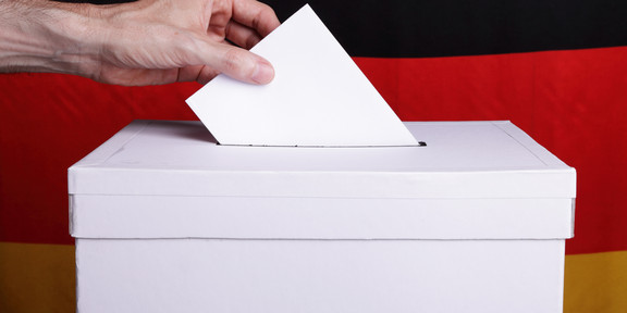Eine Person wirft einen Wahlzettel in eine Wahlurne. Im Hintergrund ist eine Deutschlandfahne