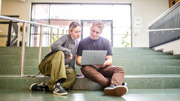 Zwei junge Erwachsene sitzen auf der Treppe im Foyer des Seminarraumgebäudes der TU Dortmund und schauen auf einen grauen Laptop.