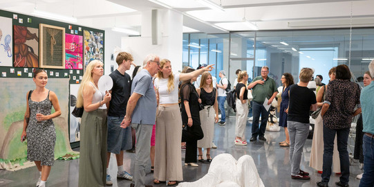 Auf dem Bild ist ein großer Ausstellungsraum zu sehen. Auf der linken Seite hängen viele verschiedene Bilder an einer Wand. Zentrum des Bildes ist eine weiße Kunstinstallation sowie mehrere Kunstinteressierte.