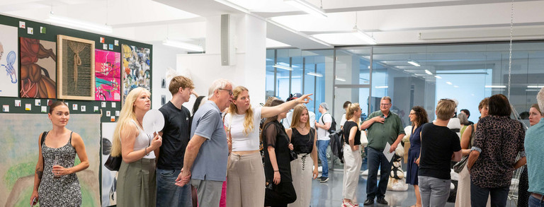 Auf dem Bild ist ein großer Ausstellungsraum zu sehen. Auf der linken Seite hängen viele verschiedene Bilder an einer Wand. Zentrum des Bildes ist eine weiße Kunstinstallation sowie mehrere Kunstinteressierte.