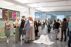 Auf dem Bild ist ein großer Ausstellungsraum zu sehen. Auf der linken Seite hängen viele verschiedene Bilder an einer Wand. Zentrum des Bildes ist eine weiße Kunstinstallation sowie mehrere Kunstinteressierte.