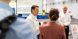 A man, Hubertus Heil, speaks through a microphone to a group of people. The group is standing in a large hall, something is projected on a screen in the background.