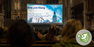 Das Foto wurde von den Kirchenbänken aus aufgenommen. Im Fokus steht eine Leinwand und ein Mann, der an das Publikum gewandt spricht.