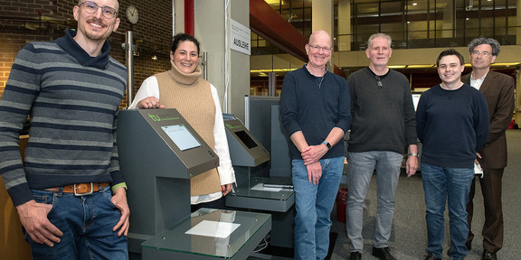 Six people stand next to new book lending equipment in a library.
