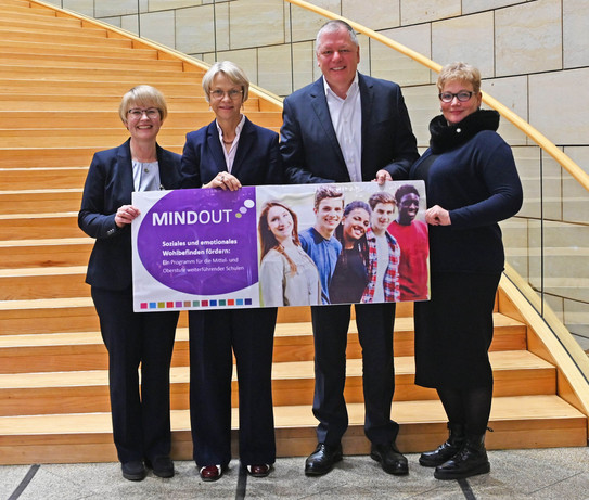 Vier Personen stehen vor einer großen, hölzernen Treppe und halten ein Plakat mit der Aufschrift "MindOut"