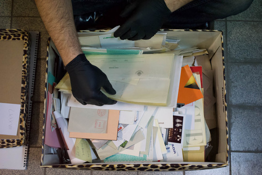 Two hands with black gloves sort through a box filled with very different documents.