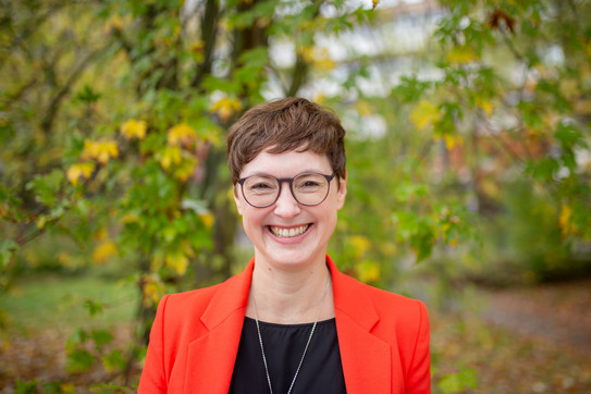 Porträtfoto: Ein Porträtfoto von Prof. Julia Faisst vor Bäumen auf dem Campus Nord. Julia Faisst hat kurze braune Haare und trägt eine Brille und einen orangen Blazer.