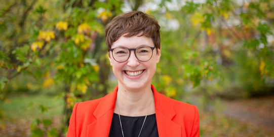 Porträtfoto: Ein Porträtfoto von Prof. Julia Faisst vor Bäumen auf dem Campus Nord. Julia Faisst hat kurze braune Haare und trägt eine Brille und einen orangen Blazer.
