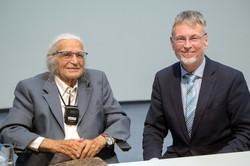Links Horst Selbiger, ein älterer Mann mit grauem Zopf und Brille, der einen grauen Anzug trägt und im Rollstuhl sitzt. Rechts Prof. Egbert Ballhorn, ein Mann mit kurzen, grauen Haaren und Brille, der einen dunkelblauen Anzug trägt und auf einem Stuhl sitzt. Beide lächeln.