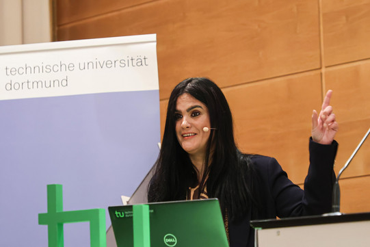 A young woman with long black hair stands at a lectern and gestures with her left hand.