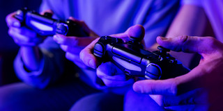 Das mit Blaufilter versehene Foto zeigt zwei von zwei sitzenden Personen gehaltene Controller