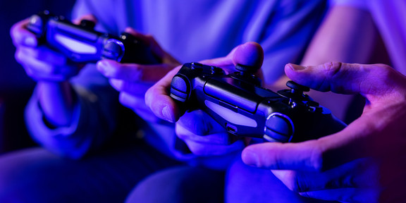 AdobeStock_416547029 Das mit Blaufilter versehene Foto zeigt zwei von zwei sitzenden Personen gehaltene Controller
