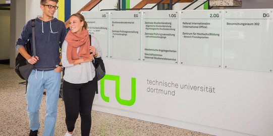 Ein Studentenpärchen läuft an einem TU-Logo im Foyer eines Gebäudes vorbei.