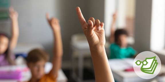Ein erhobener Finger in einem Klassenraum. Im Hintergrund sieht man verschwommen weitere Kinder.