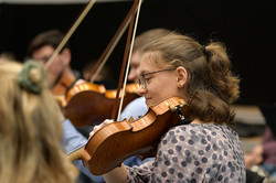 Das Bild zeigt von der Seite eine Frau, die auf einer Geige spielt. Im Hintergrund sieht man verschwommen mehrere Personen, die ebenfalls Streichinstrumente spielen.