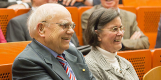 Ein älterer Mann und eine ältere Frau sitzen in einem Hörsaal und schauen lächelnd nach vorne