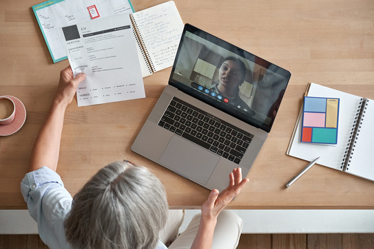 Eine Frau sitzt mit einem Laptop an einem Tisch und führt einen Videoanruf mit einer weiteren Frau. In ihrer Hand hält die Frau vor dem Laptop einen Lebenslauf.