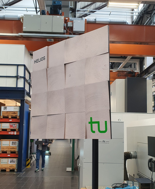 Foto: Eine quadratische Holzstruktur mit der Aufschrift „HELIOS“ und dem grünen TU-Logo steht in einer Werkhalle.