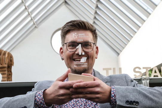 TU Sieger Junge Spitzenforschung - Siegerfoto Platz 1 Auf dem Porträtfoto sieht man den erstplatzierten Mann, der vor seinem Gesicht den ersten Preis in der Hand hält.