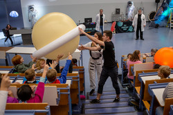 Zwei Personen geben einen großen Wasserball, der den Planeten Saturn symbolisiert, an Kinder weiter, welche in einem vollbesetzten Hörsaal sitzen.