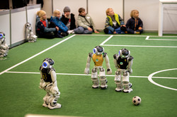 Das Bild zeigt ein kleines Fußballfeld. Am Rand sitzen Kinder und an der Mittellinie stehen drei Roboter, die ihren Blick auf den Fußball richten.
