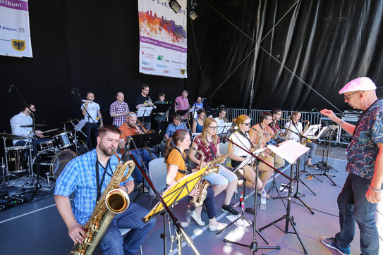 Musicians in summery, colorful outfits sit and stand on a stage playing their instruments, in front of them stands a conductor