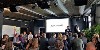 Launch "IP-Transfer 3.0" Eine Gruppe von Menschen sitzt auf Stühlen vor einer Bühne mit Leinwand, auf der das Wort "SPRIN-D" steht. Auf der Bühne steht eine Frau.