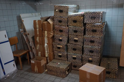 In a white-tiled art cellar, there is a large pile of different cardboard boxes in one corner. Many of the boxes have a leopard pattern.