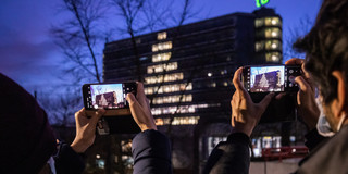 Zwei Personen fotografieren mit ihren Smartphones den Mathetower, dessen Fenster so beleuchtet sind, dass die Silhouette eines Tannenbaums zu erkennen ist.